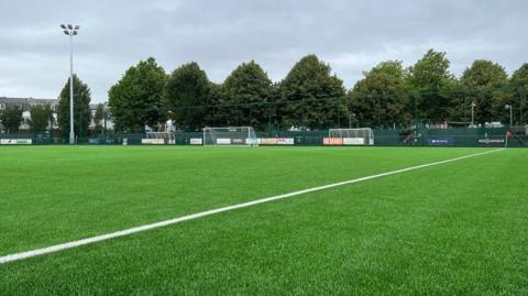 A green sports pitch with goal nets in the background.