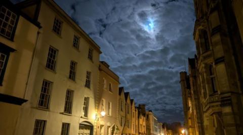 A street runs down the centre of the photo with tall stone buildings on either side. Overhead thick clouds part in just a single spot where the moon can be seen. 