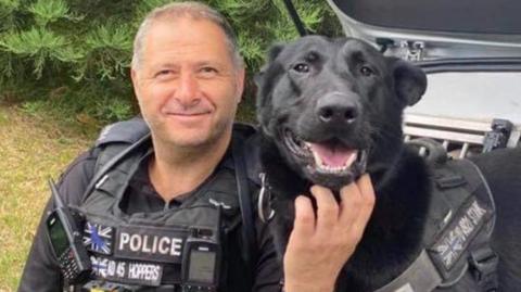 A man with short grey hair and wearing a black police officer's uniform, smiles as he sits on the back of a car. A black-coloured dog is next to him. The man is scratching the dog under his chin.