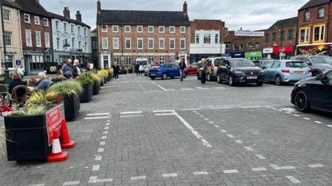 A view of a large market place, laid with grey block paving stones, with parking in the centre and black planters, topped with green shrubs, down one side, separating the road from a pedestrian area. Cars are parked to the right, while people sit at al-fresco dining tables to the left. The square is surrounded by buildings, some Georgian-style, some more modern retail units.