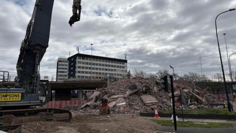 A digger in the foreground on the left-hand side, next to a pile of rubble where Computer House once stood. Behind it is the doomed Gateshead Flyover and another large office block on the other side of the raised road.