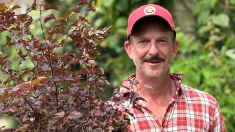 A man in a red and white checked shirt and red baseball cap holds a purple plant. 