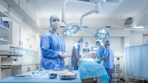 A nurse dressed head to toe in blue scrubs including a blue face mask and hair net. She is standing in front of a table with surgical equipment on it, inside an operating theatre. In the background, a patient lies on a bed under a blue blanket, with three other healthcare workers around them, also in scrubs.