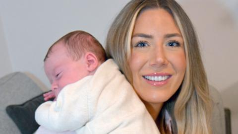  Grace Bell holding her baby son Hugo Powell. Grace is facing the camera and has centre parted blonde hair. Hugo is wearing a cream outfit and is held on her shoulder