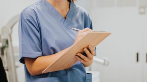 A young nurse is wearing blue scrubs and is taking notes down on a light wooden clipboard.