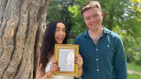A man and a woman stand next to a tree in a park, with foliage behind. The woman, on the left, has long black hair and holds up a piece of text in a 7x6 golden frame. On her right,  the brown-haired man wears a dark blue shirt with long sleeves. Both are smiling.