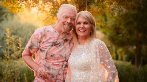 Against a leafy sunlit backdrop, Sir Richard Branson smiles at the camera, one hand holding a champagne flute, the other around Joan Templeman, his wife. Branson wears a t-shirt printed with her face. Templeman has shoulder length blonde hair with a die fringe, and smiles at the camera. She wears a white sparkly dress with a bell sleeves in netting, studded with silvery stars. The occasion was her birthday this year.