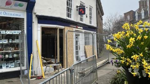 A view of the exterior of a Nationwide that has been damaged during a ram-raid incident. The front window has been removed and some cardboard blocks cover part of the gap. Metal fences surround the building. A bunch of yellow daffodils can be seen to the right of the image.