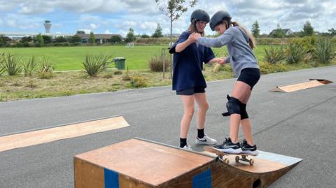 Two individuals are on a paved path in an outdoor setting with greenery and cloudy skies. One person is standing on a skateboard at the top of a small wooden ramp, while the other is assisting by holding their hands. Both are wearing helmets, and one is also wearing knee pads