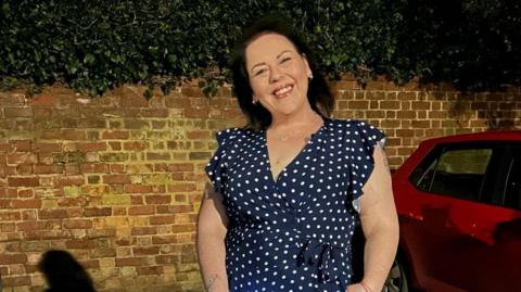 Stephanie is standing in front on a brick external wall with a red car on the right hand side. She has black shoulder-length hair and is wearing a blue short sleeve dress with white polka dots on it. 