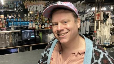 Mike Baxter, a man in a peach shirt and blue and brown leopard print cardigan. He is looking at the camera and smiling and is wearing a cap. 