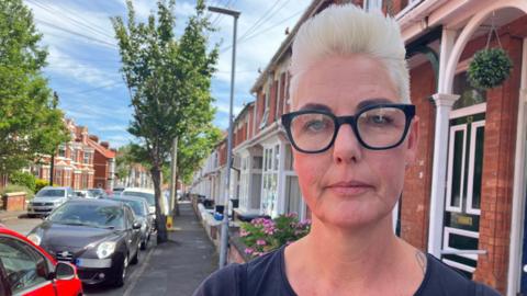 Lucy Pearce is looking at the camera while standing on a street lined with red-brick terraced homes. She has bleached blonde short hair and is wearing large black-rimmed glasses. 