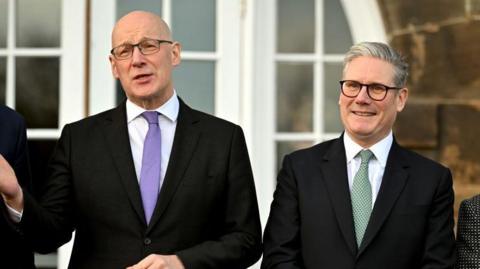 Sir Keir Starmer, with grey hair and  glasses, stands to the right of John Swinney, bald with glasses. He has his left hand raised in front of him with an open palm. Both are wearing dark suits and white shirts. Starmer is wearing a green tie while Swinney is wearing a purple one. 