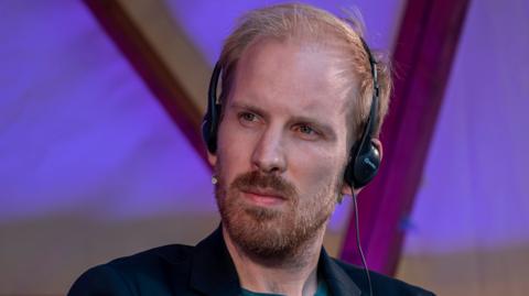 Rutger Bregman, wearing headphones, is looking to his right with a thoughtful expression - he is wearing a dark suit that can just be seen, he has blond-red hair and a short beard and moustache - he is inside a building with purple lighting behind him 