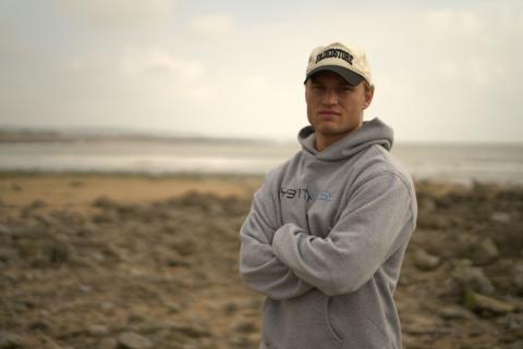 Hari Morgan standing with his arms crossed, wearing a cap and grey hoodie on the beach.