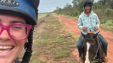 A selfie taken by a woman who smiles while wearing a helmet and pink glasses. A man in the background smiles while riding a horse. A red dirt and grassy field is in the background. 