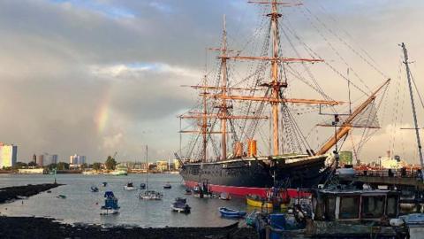 A grey sky sits behind HMS Warrior, Britain’s first iron-hulled, armoured battleship. To its left there are a number of smaller vessels bobbing in the sea with a rainbow just about visible through the clouds.