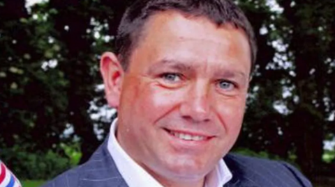 Close-up of a smiling, tanned man in a grey suit jacket and white collar shirt