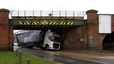 A lorry with a white cab and blue wagon appears wedged under a low railway bridge, which has red-brick walls and yellow-and-black chevrons along the cross-beam.