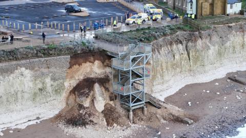 A collapsed cliff. Debris has surrounded a metal staircase.