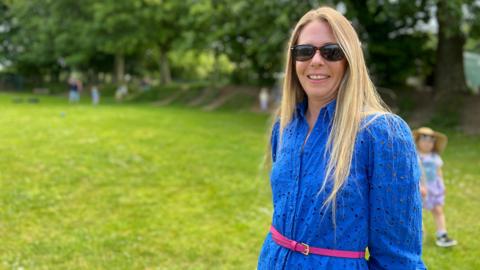 Laura has long blonde hair stands in a grassy park area, wearing a bright blue dress with a pink belt. The person's face is blurred for privacy. In the background, there are trees and other people, including a child wearing a hat. The sky is partly cloudy