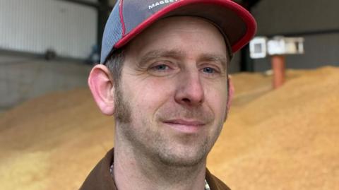 Farmer Philip Weston stands in a barn full of grain. he has sideburns and wears a blue baseball cap.