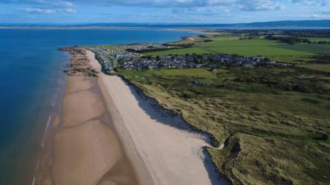 An aerial view of the village of Embo and a nearby holiday park on a beautiful sunny day. The village is small and is on the Moray Firth coast. There is a sandy beach and grass-covered sand dunes near to it.