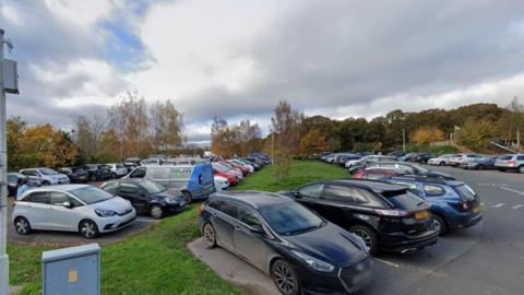 A large car park that is full of cars. There is a small strip of grassland in between two sections of the car park with trees in the distance. 