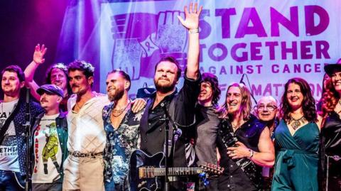 Fourteen musicians gathered together in the centre of a stage lit with pink lights to hank thank the crowd at the end of a show.