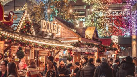 Broadgate, Coventry city centre. Christmas decorations and lights are everywhere. People are ordering food from stalls. It is winter and they have warm clothes on. 