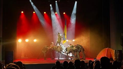 On stage actors in military fatigues hold a Ukrainian flag under spotlights. The heads of audience members can be seen in the foreground.