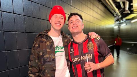 Two men stand in a black corridor. The man on the left wears a camo jacket with  a white T-shirt underneath. He also wears a red woollen hat. The man on the right has dark hair and wears a red and black horizontally striped football top. In his left hand he holds a glass.