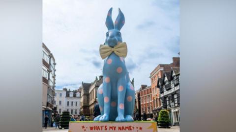 A large blue statue of a rabbit with pink spots on a stand which reads What's my name and a number of tall buildings behind it
