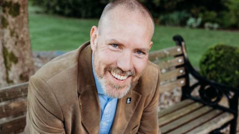 Graham, a man with a bright smile and grey stubble, who is wearing a brown jacket and blue shirt and sitting on a bench.