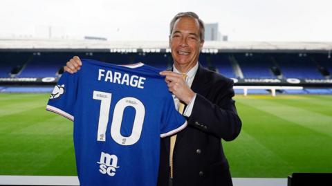 Nigel Farage poses with a blue Ipswich Town home shirt with 'FARAGE 10' on the back. He is standing next to the pitch at the Portman Road stadium.