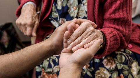 An older person is being comforted by somebody else who is holding their hands from outside the shot.