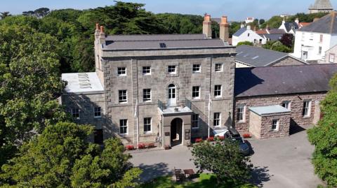 A drone shot of the Alderney government building. It is a classic-looking grey brick building with small windows and a large porch entrance. It is surrounded by trees.
