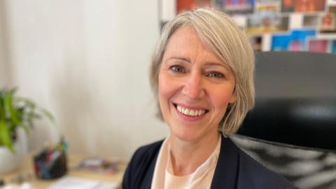 Caroline Santer, headteacher of The King's School in her office. She has bobbed blonde hair and is wearing a smart blue jacket. She is smiling towards the camera.