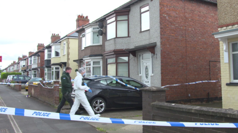 A person in a white overall suit walks intwo a semi-detahced home wiht large bay windows. Police tape is set up around the building.