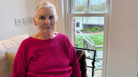 A woman wearing a pink jumper sits on a cream sofa. A garden and bench can be seen from the window behind her.