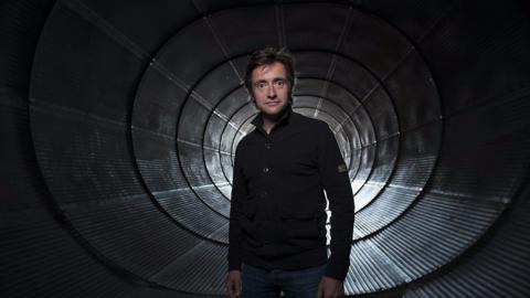 Presenter Richard Hammond dressed all in black and standing silhouetted in a wind tunnel.