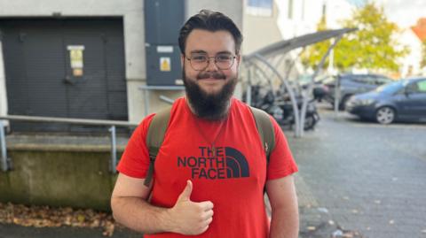 A young man with black hair, glasses and a beard wearing a red t-shirt smiles at the camera