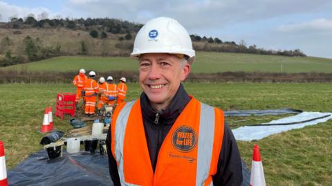 Joff Edervane wearing an orange Southern Water high vis vest and white hard hat smiling at the camera. Joff is standing in a field where the work is happening and behind him are five workers in the same clothes as him and there are tarpaulin sheets on the floor. The field is in the crest of a valley between two hills, one of which can be seen in the background