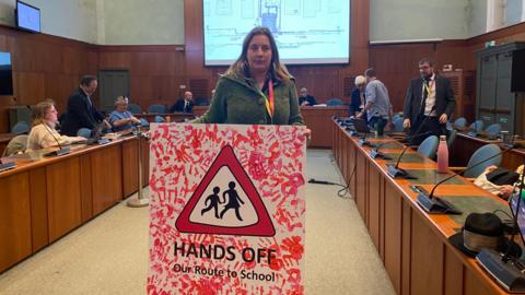 Bridget Petty holds a large cardboard sign with a pedestrian road sign on it showing an adult and child holding hands as they walk in a red-lined triangle. The sign says 'Hands off our route to school' and is covered with bright red handprints of children. She is holding the sign in the middle of the council chamber, with people taking their seats. There is a large display screen behind her showing plans. 