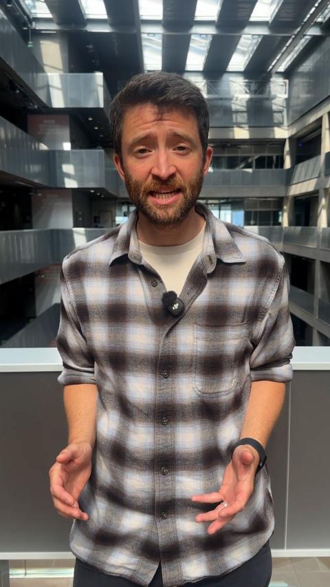A man with brown hair and a checked shirt is gesturing with his hands.