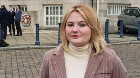 A woman in a pink blazer with pinstripes over a white jumper. She is stood outside a large stone building and has a stern expression.