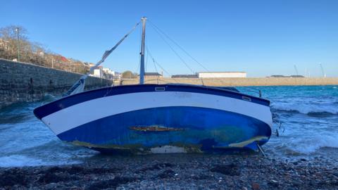 A boat washed up along a rocky bay. The yacht is blue with a white stripe along it. There is a whole on the bottom of the vessel that looks like a rip and the sail has been completely ripped off. 