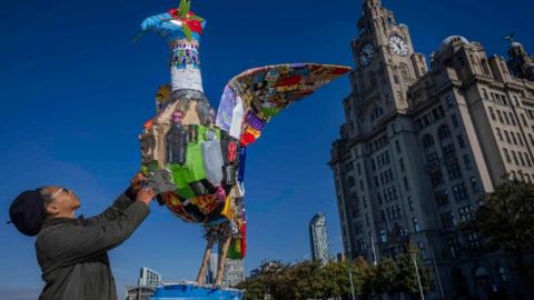 Faith Bebbington reaches towards a multi-coloured sculpture of a Liver Bird made from plastic bottles, cardboard and boxes. The Liver building is in the backdrop on a sunny day with bright blue skies.