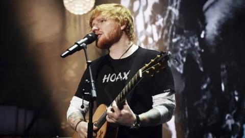 Ed Sheeran plays his guitar and sings into a microphone on a stage. He has ginger hair and a beard and is wearing a black t-shirt which says 'hoax'. 