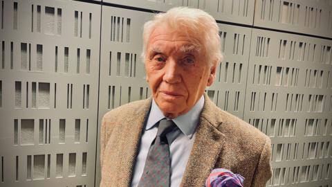 Don McCullin, wearing a light brown jacket and tie, standing in front of a grey wall with rectangular shapes on it